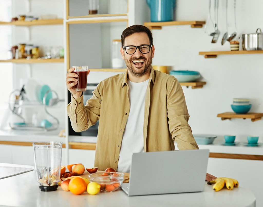 Ethan Carter holding a glass of a freshly made smoothie and smiling, with his laptop open on the kitchen counter.