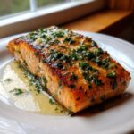 A perfectly cooked fillet of air fryer salmon topped with melted garlic herb butter, sitting on a white plate.