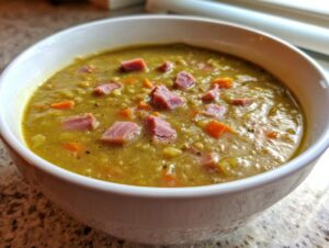 Close-up of a white bowl filled with thick, green pea soup topped with diced ham and carrots.
