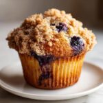 A close-up shot of a perfectly baked blueberry muffin topped with golden brown streusel, resting on a small white plate.