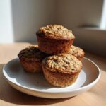 Four golden-brown banana protein muffins stacked on a white plate, lit by natural sunlight.
