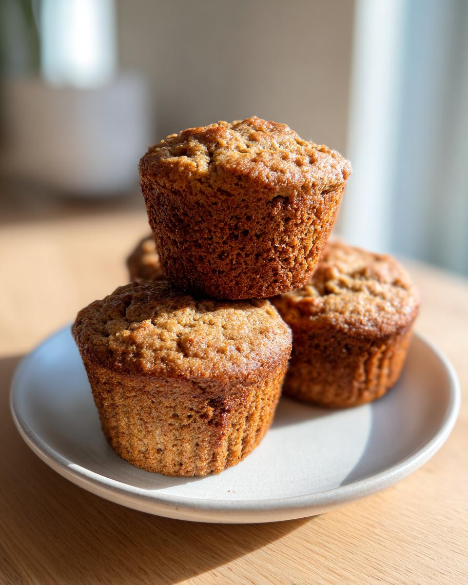 A stack of three golden-brown banana protein muffins resting on a light gray plate in bright sunlight.