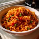 Close-up of a white bowl filled with fluffy, orange-red spanish rice mixed with bright green peas and diced carrots.