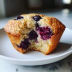 A close-up of a moist blueberry protein muffins sitting on a white plate, showing the vibrant purple filling where a bite has been taken.