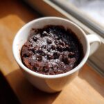 A rich chocolate mug cake topped with melted chocolate chips and powdered sugar, sitting in a white mug.