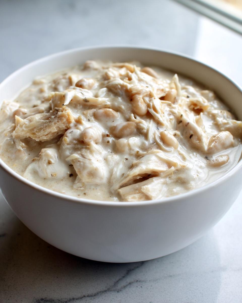 A close-up of a white bowl filled with creamy crockpot white chicken chili, showing shredded chicken and white beans.