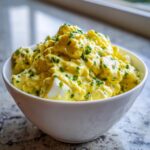 A close-up of a generous serving of creamy, yellow egg salad recipe topped with fresh green chives in a white bowl.