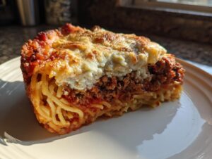 A close-up slice of baked spaghetti pie showing layers of pasta, meat sauce, and a creamy cheese topping.