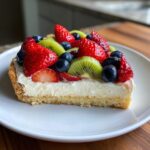 A cross-section slice of fruit pizza sugar cookie topped with cream cheese frosting and fresh strawberries, blueberries, and kiwi.