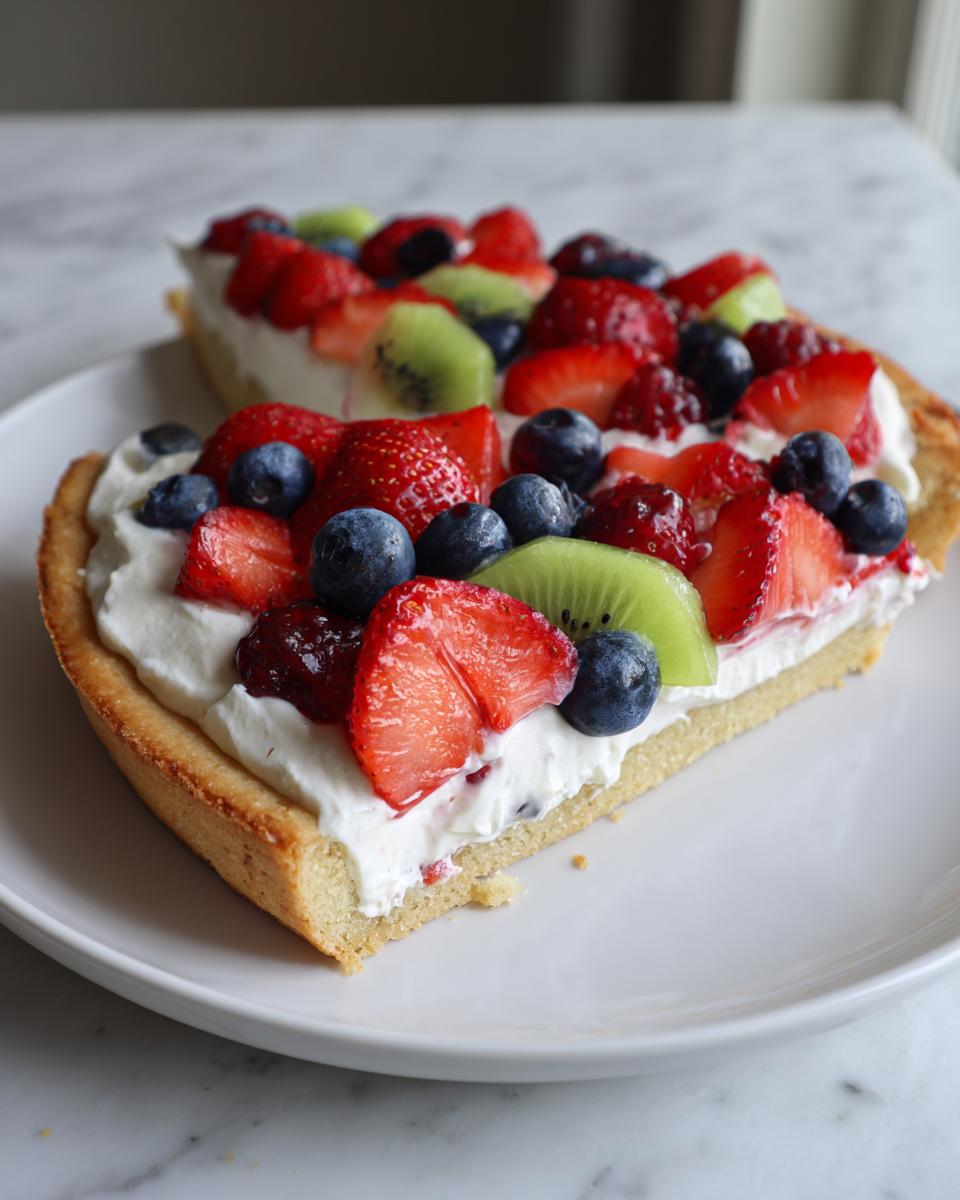 A slice of fruit pizza sugar cookie topped with whipped cream, strawberries, blueberries, and kiwi.