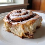 A close-up shot of a delicious, soft cinnamon roll topped with thick white icing, sitting on a white plate.