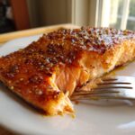 Close-up of a perfectly cooked salmon fillet with a thick, shiny glaze being flaked by a fork on a white plate.