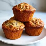 A stack of three freshly baked, golden brown banana nut muffins topped with visible walnuts on a white plate.