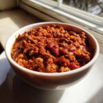 Close-up of a white bowl filled with rich, meaty crockpot chili, illuminated by sunlight near a window.