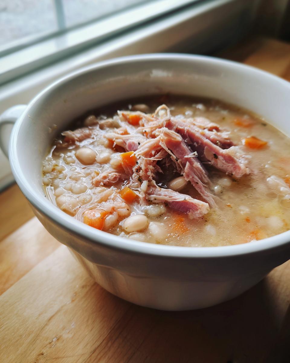 Close-up of a white bowl filled with rich Ham and bean soup, topped with shredded ham and diced carrots.