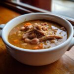 Close-up of a white bowl filled with rich Ham and bean soup, topped with shredded ham and visible carrots.