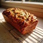 A freshly baked loaf of Italian herb and cheese bread cooling on a wire rack near a window.
