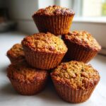 A close-up stack of five golden-brown, moist bran muffins with textured tops, set against a bright, blurred background.