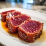 Four thick slices of seared tuna steak, rare in the center, resting in a pool of marinade on a white plate.