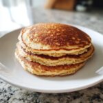 A close-up of a fluffy stack of four golden-brown almond flour pancakes on a white plate.