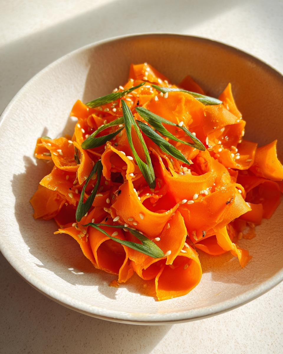 Close-up of a vibrant raw carrot salad made with shaved carrot ribbons, topped with sesame seeds and green onions.