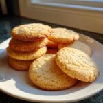 A stack and pile of golden brown 3-ingredient butter cookies resting on a white plate near a window.