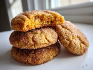 A stack of soft, orange-hued pumpkin snickerdoodle cookies coated in cinnamon sugar, with one cookie broken open.
