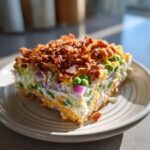 A thick, square slice of Amazing 7 layer salad showing layers of peas, red onion, cheese, and creamy dressing, topped with crumbled bacon.