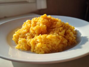 A close-up of creamy, bright yellow cheesy rice served in a mound on a white plate, catching the sunlight.