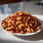 A large mound of savory, seasoned chex party mix, including Chex cereal and pretzels, piled high on a white plate.
