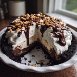 A rich, multi-layered ice cream pie with a dark crust, chocolate filling, whipped cream, fudge drizzle, and chopped peanuts.