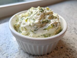 A close-up of creamy, chunky pickle dip topped with fresh dill in a small white bowl.