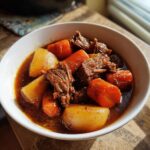 Close-up of a hearty bowl of rich pork stew featuring tender shredded meat, large chunks of carrots, and potatoes in a dark broth.