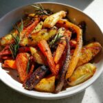 A white bowl filled with colorful roasted vegetables, including carrots and potatoes, seasoned and garnished with fresh rosemary.