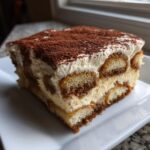 A perfect square slice of tiramisu recipe showing layers of ladyfingers and cream, topped generously with cocoa powder.