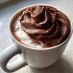 A white mug filled with hot chocolate topped with a generous swirl of rich, dark chocolate whipped hot chocolate.