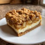 A single, decadent square of apple cheesecake bars showing a graham cracker crust, creamy cheesecake layer, and thick apple crumble topping.