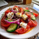 Close-up of a vibrant, authentic greek salad featuring large chunks of tomato, cucumber, red onion, Kalamata olives, and feta cheese, drizzled with dressing.