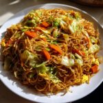 A large white plate piled high with savory, saucy fried noodles mixed with bright orange carrots and green cabbage.