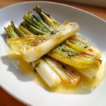 A close-up of tender, glossy braised leeks resting in a pool of buttery sauce, garnished with herbs.