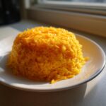 A perfectly molded mound of bright yellow cheesy rice topped with shredded cheddar cheese, sitting on a white plate.