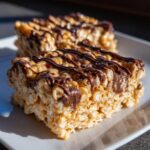 Close-up of two chewy peanut butter rice krispie treats drizzled with melted chocolate on a white plate.