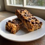 Two rich, chewy no bake granola bars topped with melted chocolate chips, resting on a white plate.