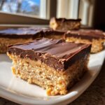 A close-up of a thick, square peanut butter bars slice with a rich chocolate topping, sitting on a white plate.