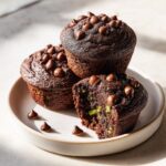 Three rich chocolate zucchini muffins stacked on a plate, one is bitten showing green zucchini pieces inside.