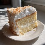 A generous slice of moist coconut poke cake topped with whipped cream and toasted coconut flakes.