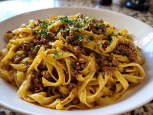 A close-up of a white bowl filled with creamy beef pasta, featuring wide egg noodles coated in a rich sauce and topped with ground beef and parsley.
