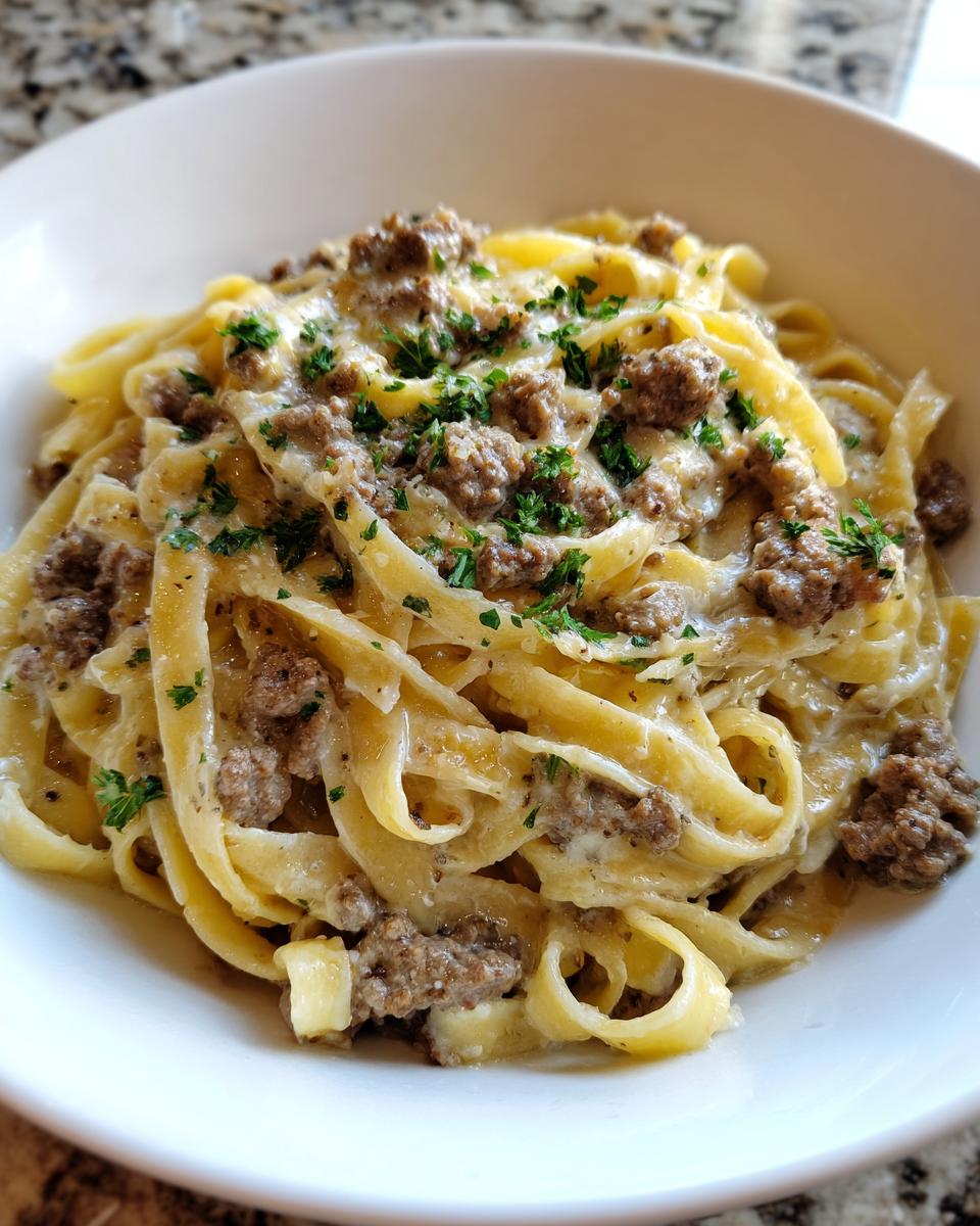A close-up of a white bowl filled with creamy beef pasta, featuring fettuccine noodles and ground beef, topped with parsley.