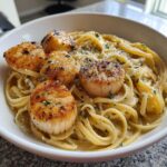 A white bowl filled with creamy seafood pasta topped with five perfectly seared scallops and sprinkled with parsley.