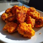 A close-up of several crispy, bright orange buffalo cauliflower bites piled on a white plate, catching bright sunlight.
